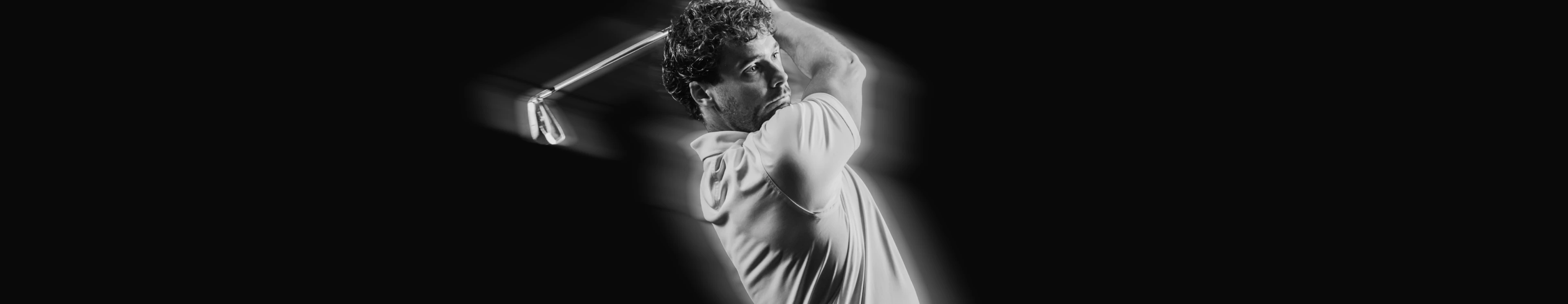 Man in a white shirt swinging a golf club with focus and intensity, captured in black and white against a dark background.