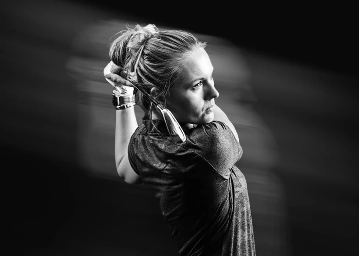 Black and white image of a woman holding a golf club in a swing motion, focused and looking into the distance.