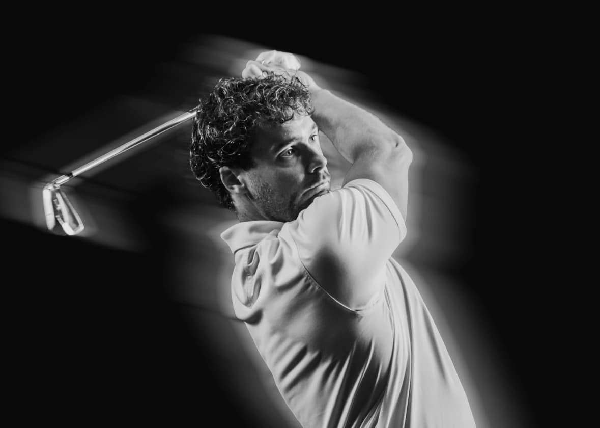 Man in a polo shirt swinging a golf club, captured in motion, with a focused expression on a dark background.
