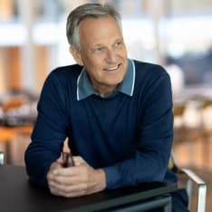 Smiling older man with gray hair in a blue sweater sitting at a table, holding a small object with both hands, in a brightly lit room.