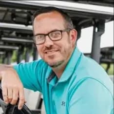 Man with glasses and short hair, wearing a teal polo shirt, smiling while sitting outdoors under a metal roof structure.